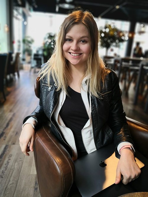 Meg: smilende ung kvinne ikledd skinnjakke, sittende i skinnstol i cafémiljø med Macen i fanget